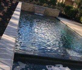 Rectangular backyard pool with rippled water and a small waterfall feature, surrounded by trees and fencing.