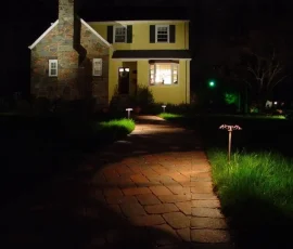 A brick walkway leads to a lit house entrance at night, bordered by illuminated path lights and green grass.