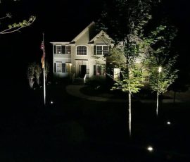Two-story house at night with exterior lights illuminating the facade, trees, and a flagpole in the front yard.