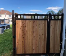 A wooden gate with black metal frame and hinges, set between a house and a grassy yard under a blue sky.