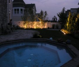 Backyard patio with a small pool, garden, and landscape lights illuminating trees and shrubs at dusk.