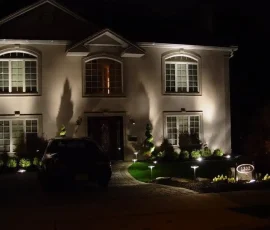 Two-story house at night with exterior lights illuminating the facade, front yard, and driveway with parked car.