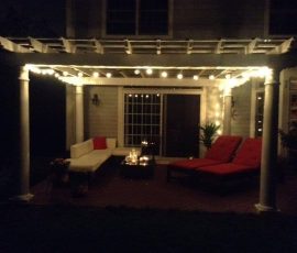 Covered patio at night with string lights, lounge chairs with red cushions, a sofa, and a small table with candles.