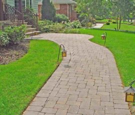 Curved stone walkway with lanterns, bordered by green grass and landscaped plants leading to a house entrance.