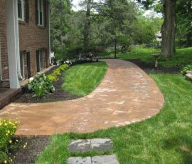 Curved brick pathway bordered by mulch and flower beds, leading through a green lawn beside a brick house.