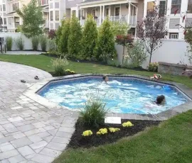 Two people are swimming in a small backyard pool surrounded by grass, plants, and patio pavers.