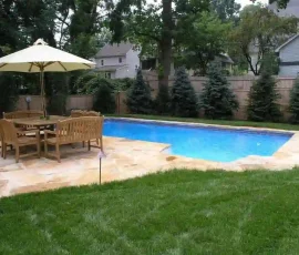 A backyard swimming pool with a patio area, wooden table and chairs, and a large umbrella, surrounded by grass and trees.