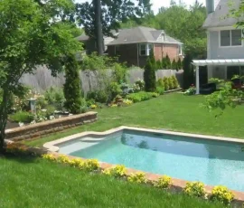 Rectangular backyard pool surrounded by grass, flowers, and landscaping with a house and patio in the background.