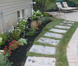 Stone pavers lead through a garden with mulch and flowers beside a pool and two lounge chairs on a patio.
