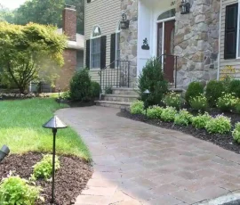 Brick walkway bordered by landscape lighting, shrubs, and flowers leads to the front steps of a stone-front house.
