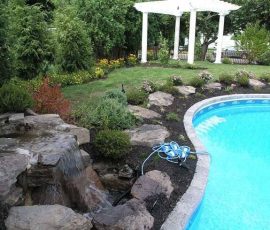 A backyard pool with a stone waterfall, garden landscaping, and a white pergola in the background.