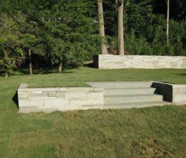Stone steps with low retaining walls lead up to a grassy lawn area surrounded by trees.